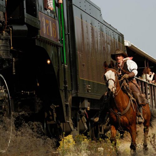 Actors on horseback dressed as Wild West outlaws ride alongside the historic 3716 steam locomotive during the Summerland Great Train Robbery , while passengers watch the action from open-air rail cars